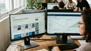 Profissional de marketing digital analisando em dois monitores as novas playlists de trilhas de SEO do Google no YouTube e métricas de queda do CAC com crescimento de vendas.